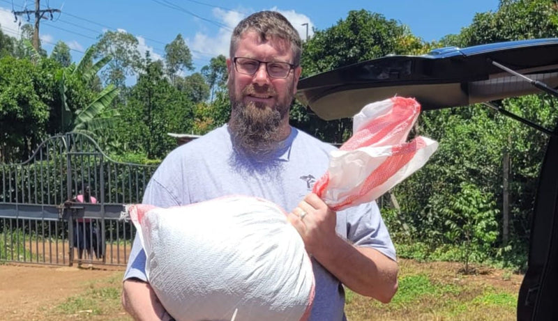 Coffee roaster Mark, a tall white man with a beard and glasses, stands in a field in Kenya holding a sack of unroasted coffee